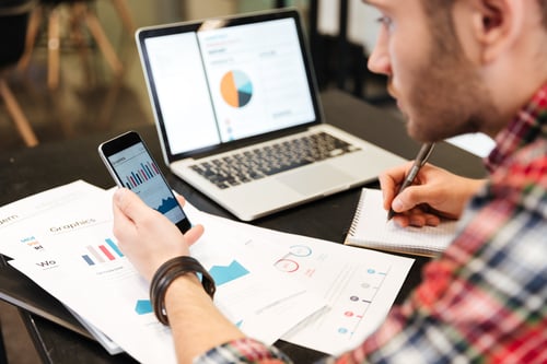 Image of a man sitting on a desk looking at graphics on his phone and papers.